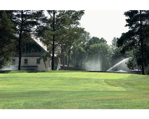 Zavlažovací systém na zeleném trávníku se stromy a budovou v pozadí