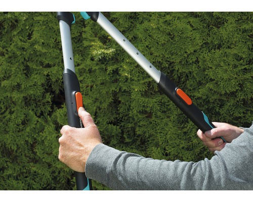 Dvě ruce ovládají zahradní nůžky před zeleným keřem.