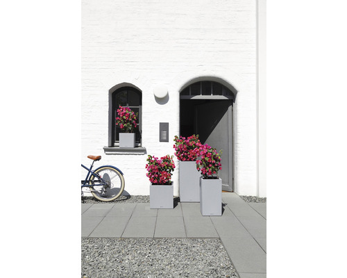 Scéna se třemi šedými truhlíky a květinami před bílou zdí domu