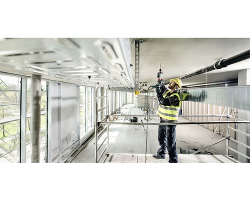 Stavební dělník na lešení pracuje s vrtacím kladivem na stropní instalaci.
