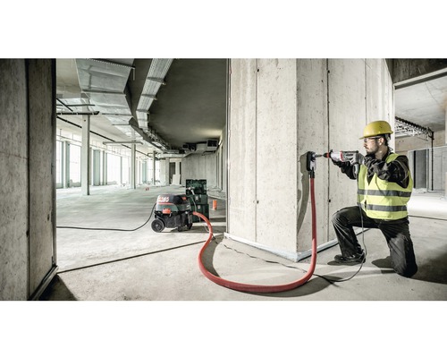 Stavební dělník vrtá s vrtacím kladivem otvor do betonové zdi, je připojeno odsávání prachu.