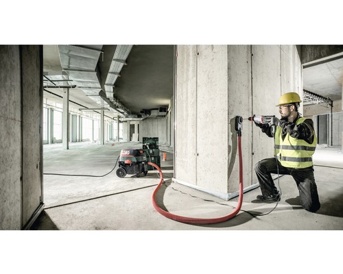 Stavební dělník vrtá vrtačkou otvor do betonové zdi. K dispozici je také průmyslový vysavač a odsávací adaptér.