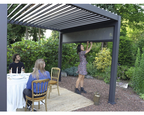 Pergola zahradní 300 x 300 cm s lamelovou střechou antracit vč. zastínění Scéna s hliníkovou pergolou s lamelovou střechou a osobou ovládající roletu, zatímco jiné osoby sedí u stolu