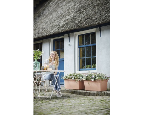 Žena sedí u stolu před domem s truhlíky, vychutnává si snídani a uvolněnou atmosféru.