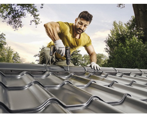 Muž připevňuje kovové střešní tašky aku šroubovákem na střechu.