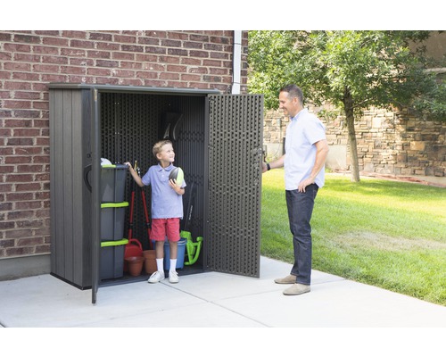 Zahradní scéna s kůlnou na nářadí, ve které stojí dítě, zatímco dospělý otevírá dveře