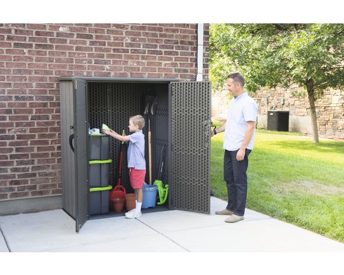 Zahradní skříň s nářadím a zahradnickými potřebami, dítě uklízí skříň, dospělý drží dveře otevřené na trávníku