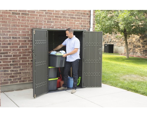 Muž ukládá zahradní nářadí do plastové zahradní skříně před cihlovou zeď