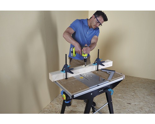 Muž vrtá vrtačkou otvor do obrobku, který je připevněn ke pracovnímu stolu pomocí šroubových svorek.