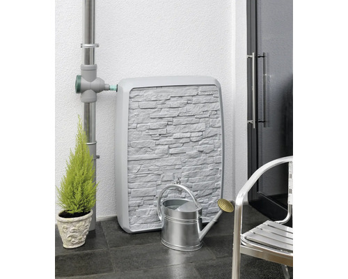 Nádrž na dešťovou vodu s kamenným vzhledem stěny, spojená se svodem, vedle ní konvička a rostlina.