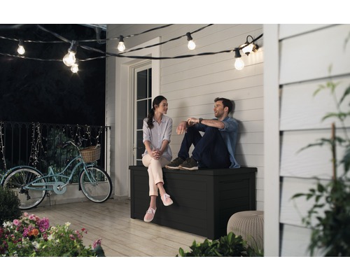 Pár sedí na černé zahradní bedně na terase pod světelným řetězem.