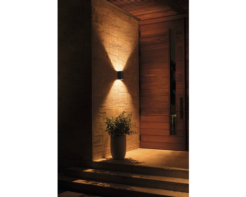 Exteriér domu s moderní nástěnnou lampou svítící nahoru a dolů, vedle dřevěných dveří a hrnkové rostliny.