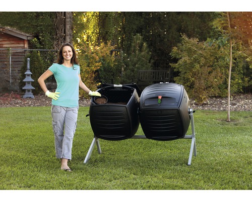 Žena stojí vedle černého dvojitého bubnového kompostéru na zahradě.