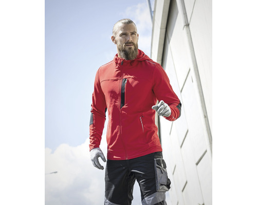 Muž s plnovousem, červená bunda s kapucí, pracovní kalhoty a pracovní rukavice před fasádou budovy