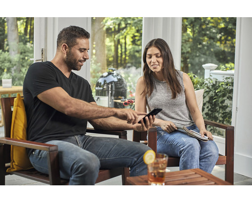 Muž a žena sedí na terase a dívají se na mobilní telefon, v pozadí kulový gril.