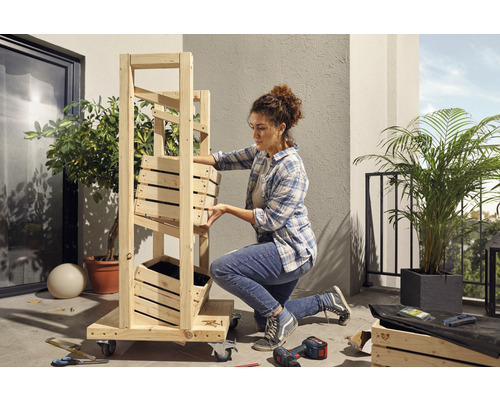 Žena montuje dřevěný regál s truhlíky na balkoně. Nářadí a rostliny jsou také vidět.