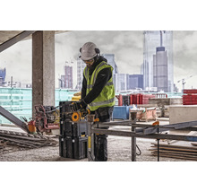 Stavební dělník s ochrannou přilbou, brýlemi, chrániči sluchu a bezpečnostní vestou pracuje s rozbrušovací pilou na staveništi.