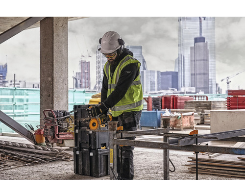 Stavební dělník s ochrannou přilbou, brýlemi, chrániči sluchu a bezpečnostní vestou pracuje s rozbrušovací pilou na staveništi.