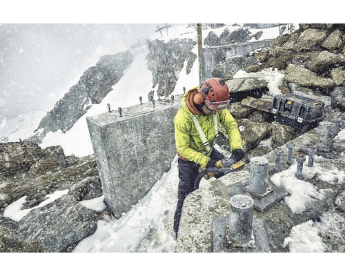 Pracovník s ochrannou helmou a chrániči sluchu pracuje s nástrojem v zasněženém prostředí. V pozadí jsou vidět hory a sníh.
