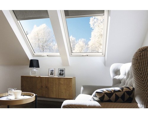 Světlá podkrovní místnost se střešními okny a roletami, zařízená komodou, lampou a křeslem