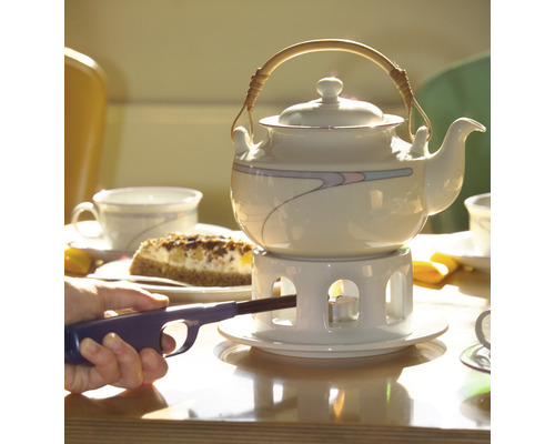 Čajová konvice na stole s dortem a osobou zapalující čajovou svíčku