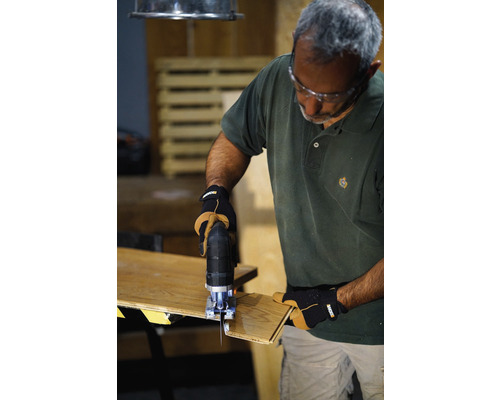 Muž používá přímočarou pilu k řezání kusu dřeva. Nosí ochranné brýle a rukavice.