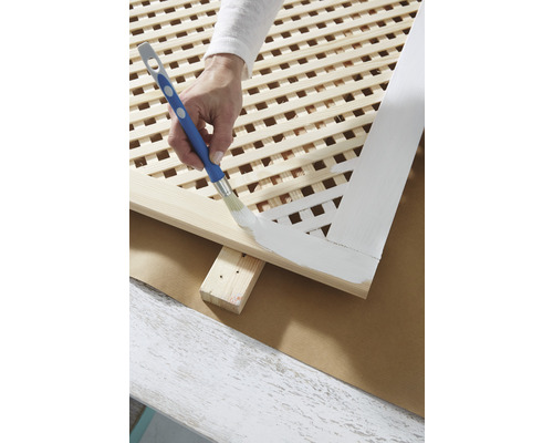 Osoba natírá dřevěnou mřížovou stěnu štětcem a barvou.