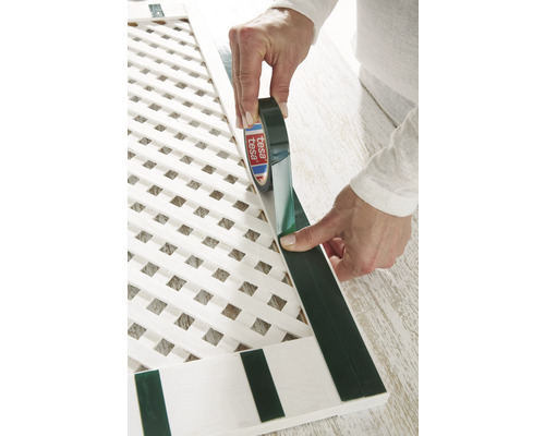 Osoba aplikuje oboustrannou lepicí pásku na bílou dřevěnou mřížkovou strukturu.