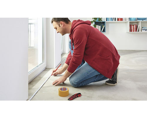 Muž měří podlahu pomocí svinovacího metru, tužky, lepicí pásky a nože.