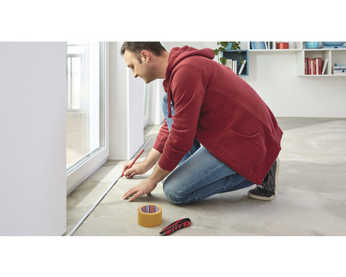 Muž měří podlahu skládacím metrem a tužkou, vedle nich leží řezací nůž a lepicí páska.