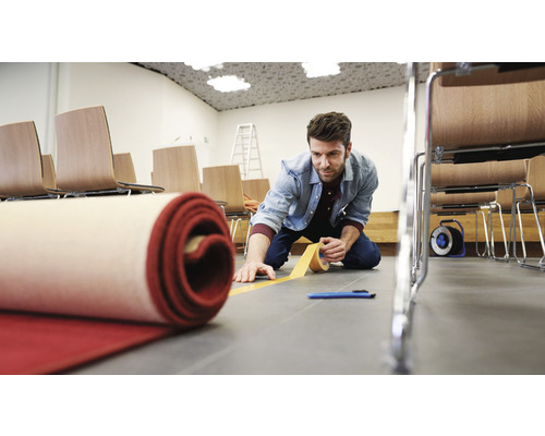 Muž pokládá koberec pomocí lepicí pásky na koberce v místnosti se židlemi.