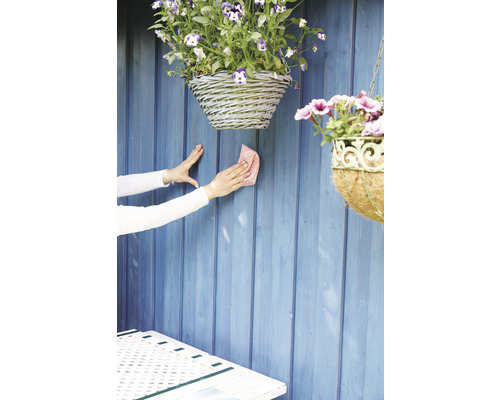 Čištění dřevěné zahradní stěny hadříkem, s visícími košíky s květinami.