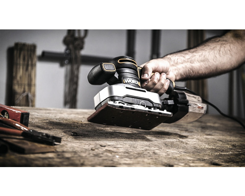 Ruka drží vibrační brusku značky Worx na dřevěném stole.