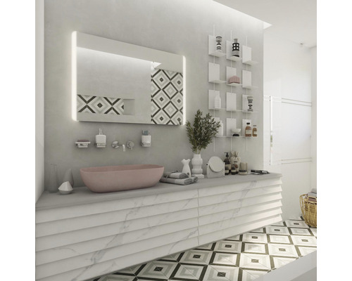 Světlá koupelna s umyvadlem, osvětleným zrcadlem, nástěnnými policemi a vzorovanou dlažbou.