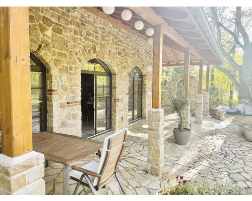 Venkovní pohled na dům s kamennou zdí, terasou, stolem a židlemi.