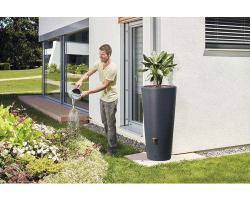 Muž zalévá rostliny na zahradě konví, v pozadí nádrž na dešťovou vodu