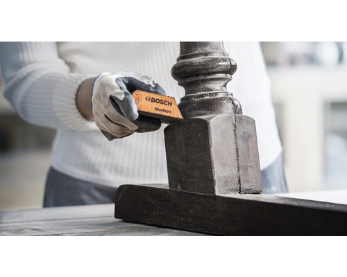 Osoba brousí tmavý dřevěný nábytek brusnou houbičkou střední zrnitosti od společnosti Bosch.