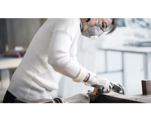 Žena brousí dřevěný obrobek brusným papírem, ochrannou maskou a rukavicemi