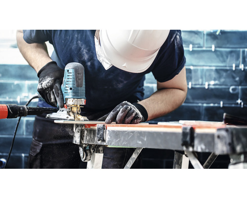 Muž s ochrannou helmou řeže obrobek na pracovním stole pomocí přímočaré pily.