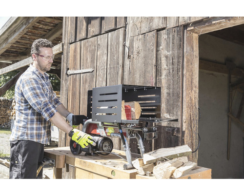 Muž obsluhuje štípačku na dřevo před dřevěnou stěnou. Štípačka stojí na pracovním stole a vedle ní leží špalky.