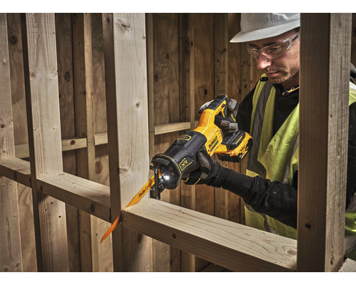 Muž s ochrannými brýlemi a ochrannou helmou řeže dřevěnou desku akumulátorovou mečovou pilou.