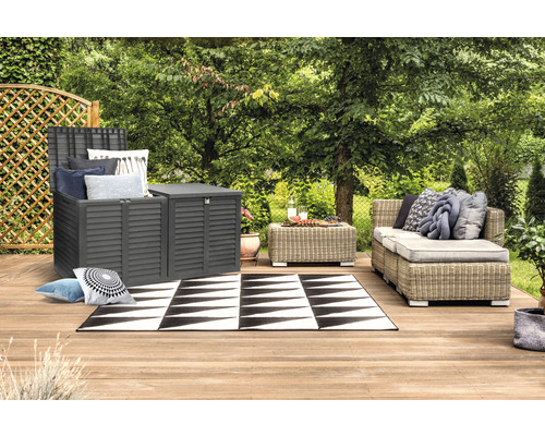 Zahradní scéna s šedou úložnou skříní, ratanovou pohovkou a černobílým kobercem na dřevěné terase.