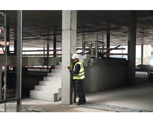 Stavební dělník měří betonový sloup svinovacím metrem v hrubé stavbě.