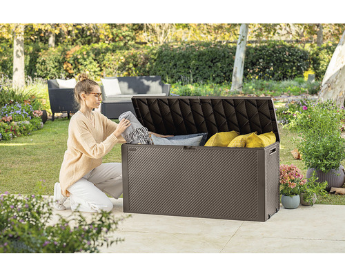 Žena ukládá deky a polštáře do hnědého zahradního boxu venku.