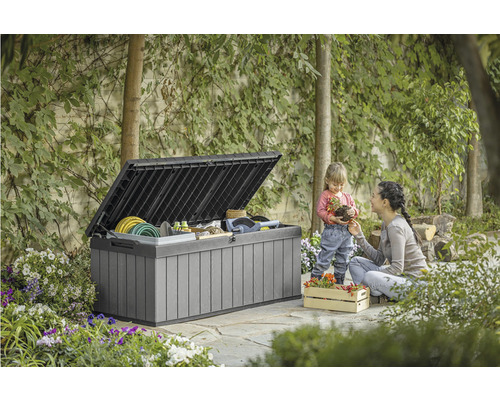 Zahradní scéna s otevřeným zahradním boxem plným zahradního nářadí a ženou s dítětem.