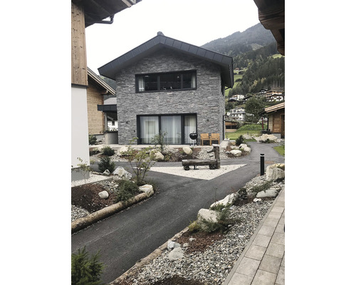 Pohled na moderní dům s dlážděnou příjezdovou cestou, skalní zahradou a grilem