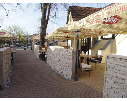 Venkovní prostor restaurace s kamennými zdmi, dřevěnou podlahou a slunečníky