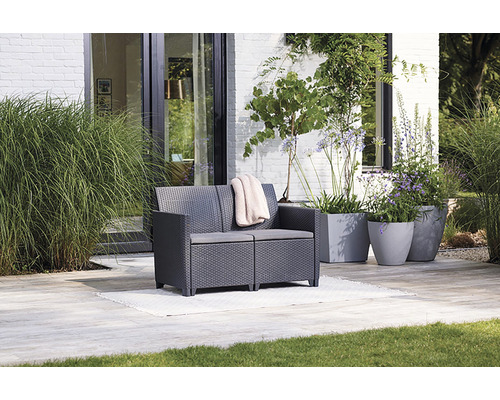 Zahrada s šedou ratanovou dvoumístnou lavicí, polštáři a rostlinami