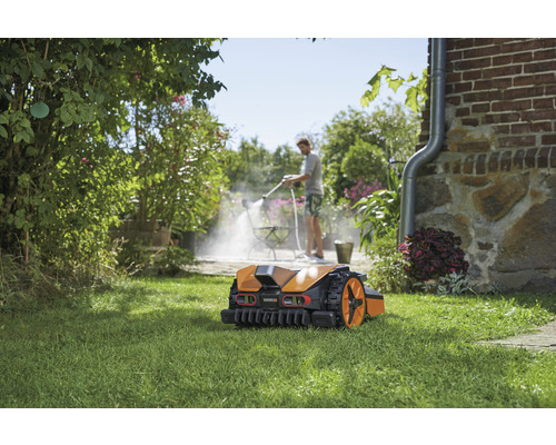 Robotická sekačka na trávníku, v pozadí osoba čistí terasu vysokotlakým čističem