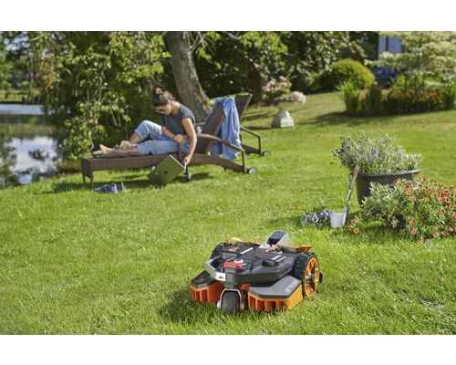 Scéna s robotickou sekačkou na zahradě, žena odpočívá na lehátku